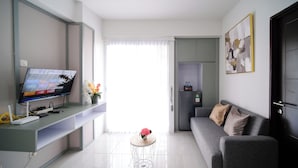 A modern living room with a flat-screen TV, a grey sofa, a small round table with a vase, and a refrigerator. - Compact And New 2Br Apartment At Suncity Residence (Sidoarjo)