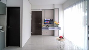 A modern living room with a dark door, a flat-screen TV on a white stand, a small round table with a flower, and a large window with sheer curtains. - Compact And New 2Br Apartment At Suncity Residence (Sidoarjo)