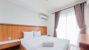 Ett hotellrum med en sänggavel i trä, en säng med vita sängkläder, en prydligt vikt handduk på sängen, ett nattduksbord och ett fönster med gardiner.