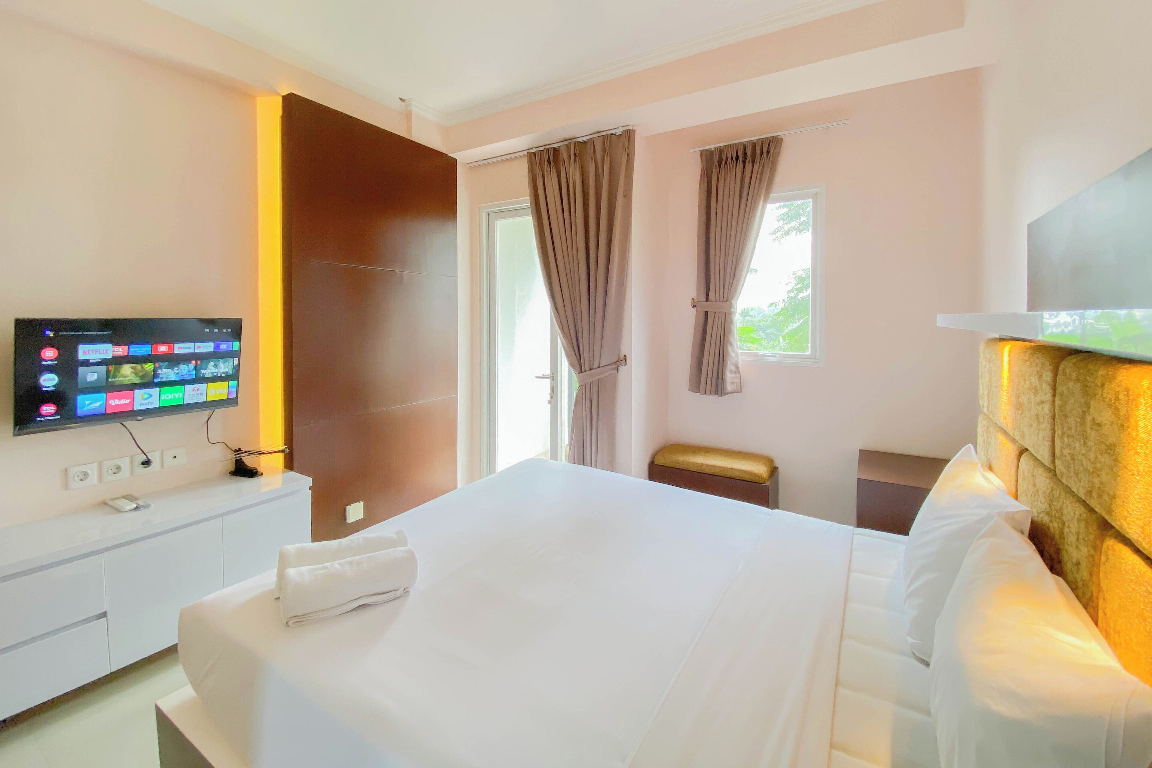 Una habitación de hotel con una cama, un televisor montado en la pared, una ventana con cortinas y una mesita de noche.