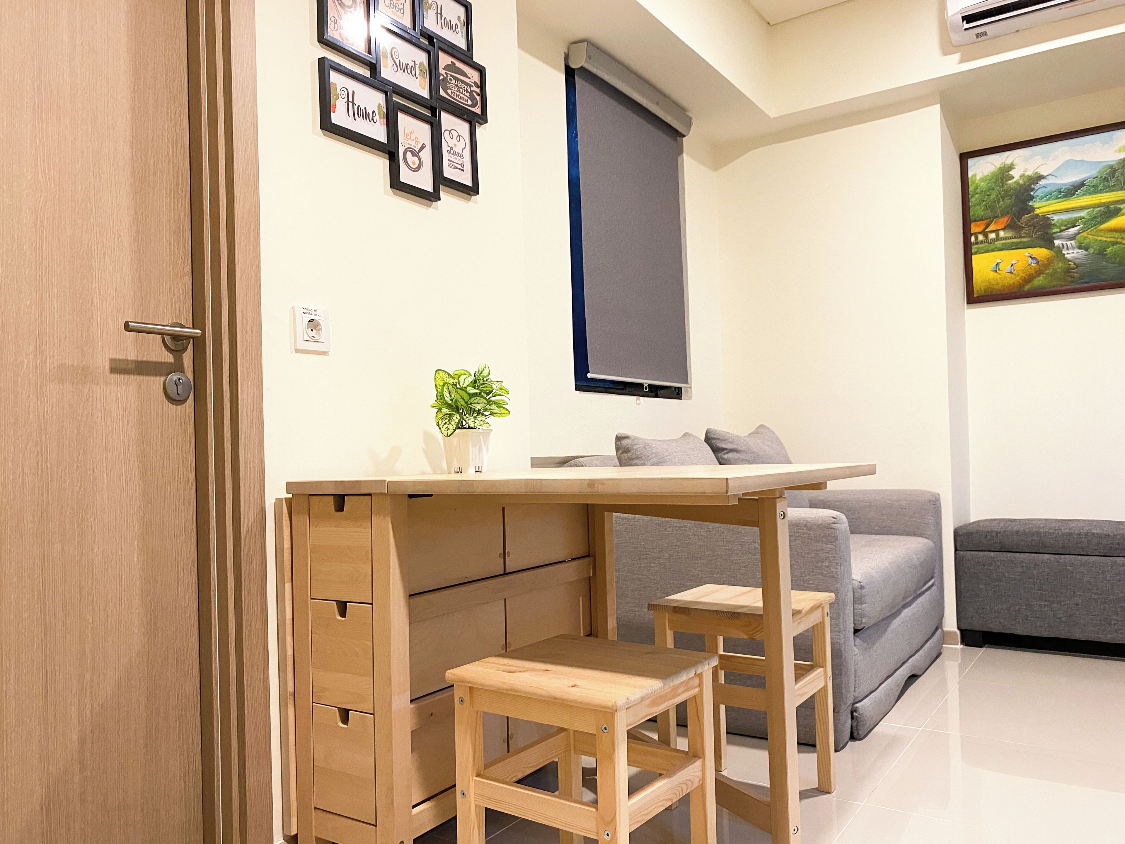 A compact living space with a wooden desk, two stools, a grey sofa, a small potted plant, and a wall-mounted TV.