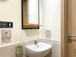 A bathroom with a sink, mirror, and two outlets, including one with a protective cover, and a waste bin.