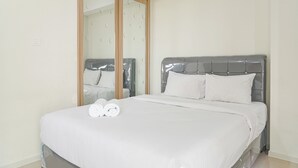 A neatly made bed with white linens and two rolled towels, a grey upholstered headboard, and a large mirror with a wooden frame.