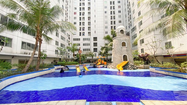 A swimming pool with slides and a water play area surrounded by palm trees and a multi-story building. - Strategic And Warm Studio At Bassura City Apartment (Jakarta)