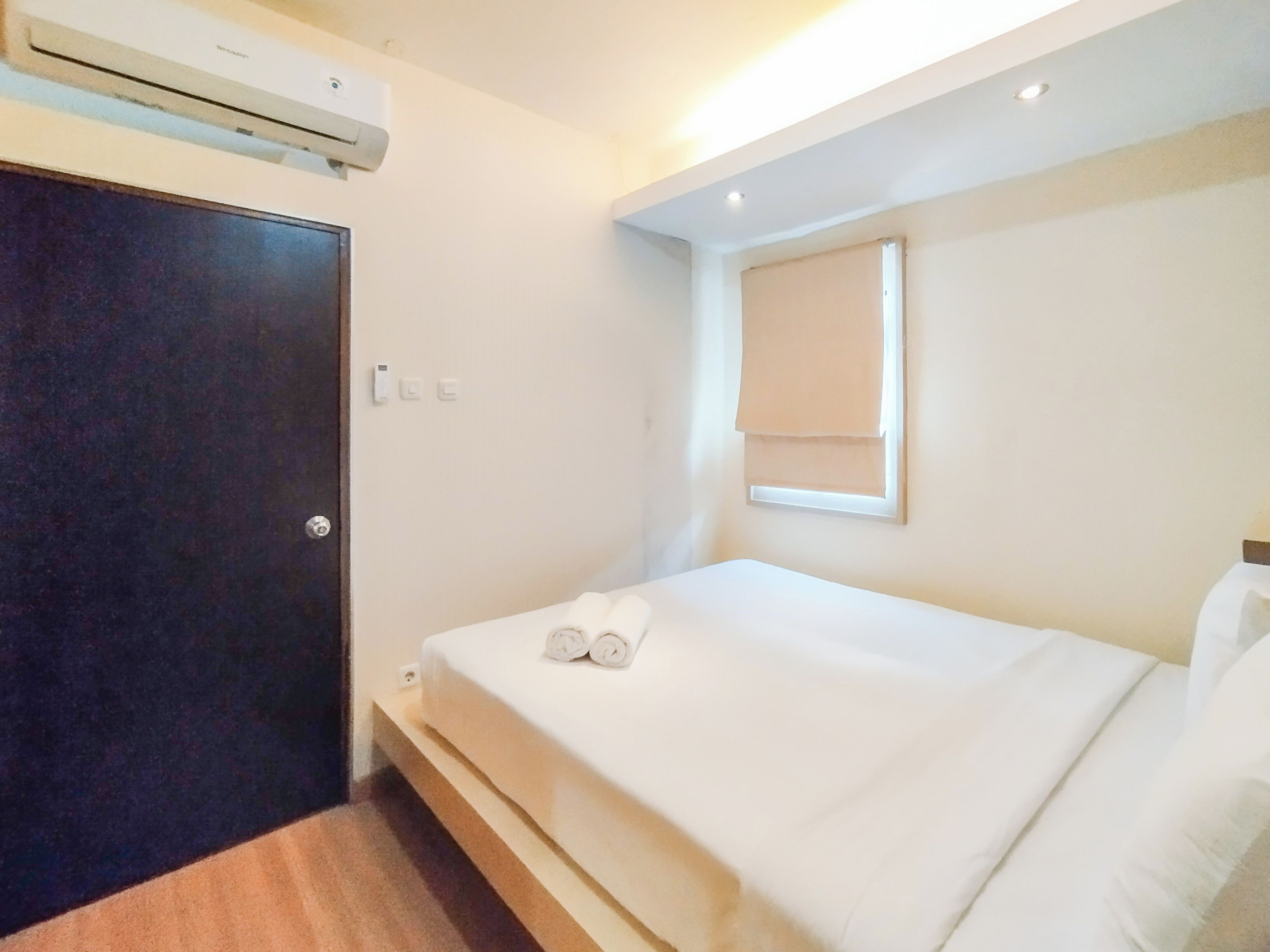A hotel room with a bed, a window with a blind, an air conditioning unit, and a door.