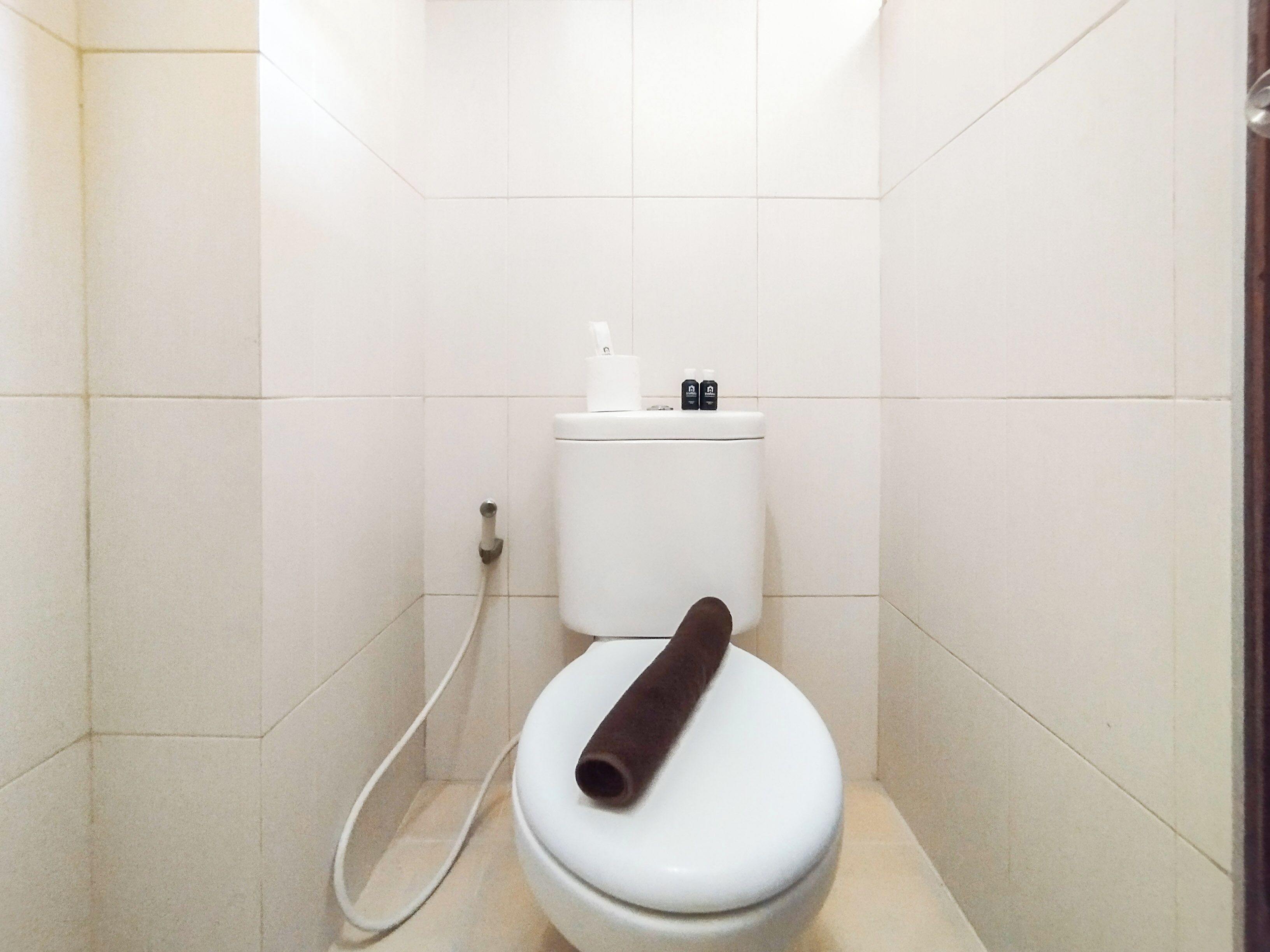 Un baño pequeño, con azulejos blancos, que incluye un inodoro, un dispensador de papel higiénico blanco y un cepillo para inodoro blanco.