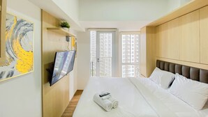 A hotel room with a bed, a TV mounted on the wall, a window with a city view, and a painting on the wall. - Cozy Designed Studio Tokyo Riverside Pik 2 Apartment (Jakarta)