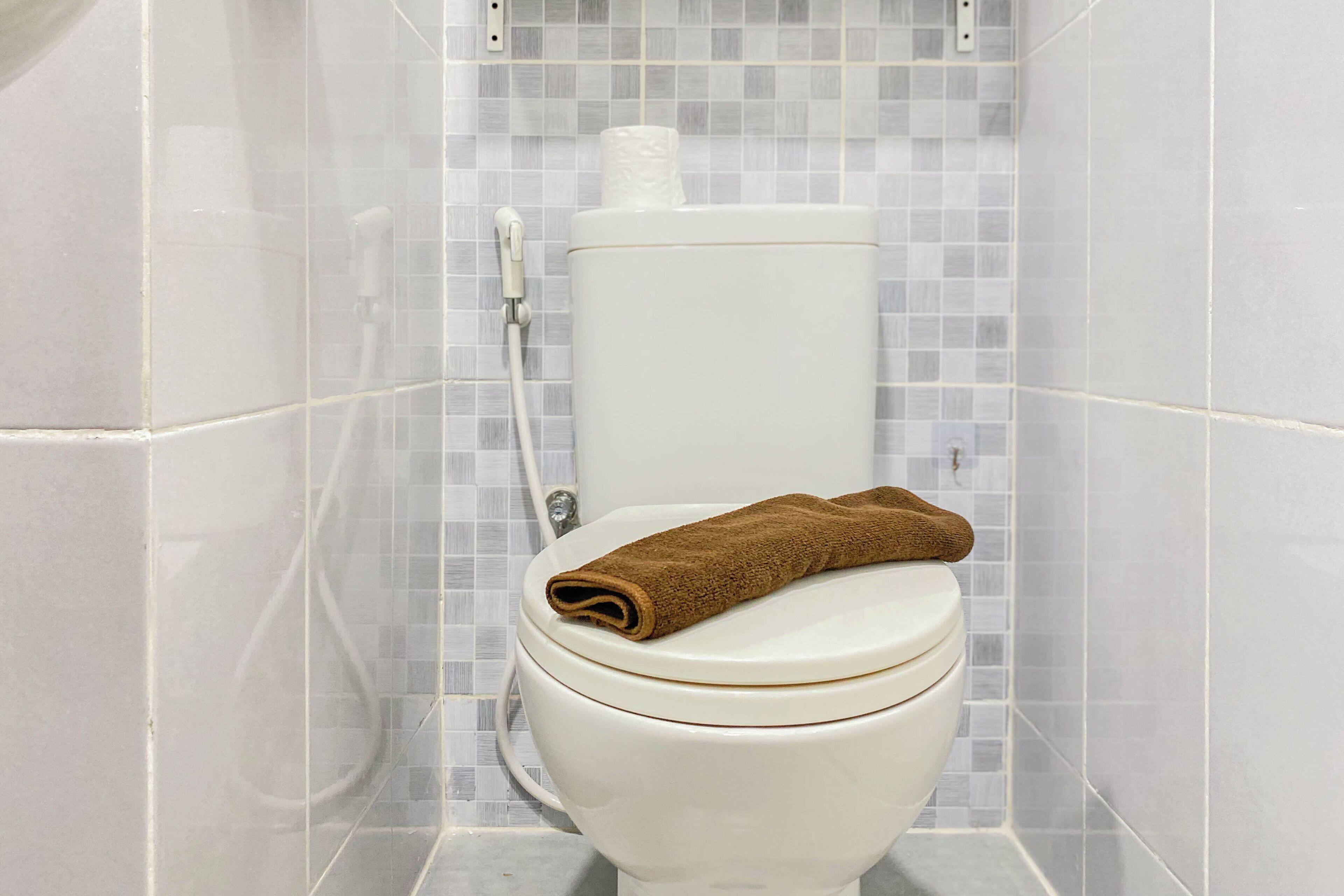 A white toilet with a brown towel on top, a roll of toilet paper, and a shower hose.