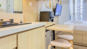 A modern hotel room with a kitchenette, a bed with neatly made white linens, a small wooden stool, and a television mounted on the wall. - Cozy Designed Studio Tokyo Riverside Pik 2 Apartment (Jakarta)