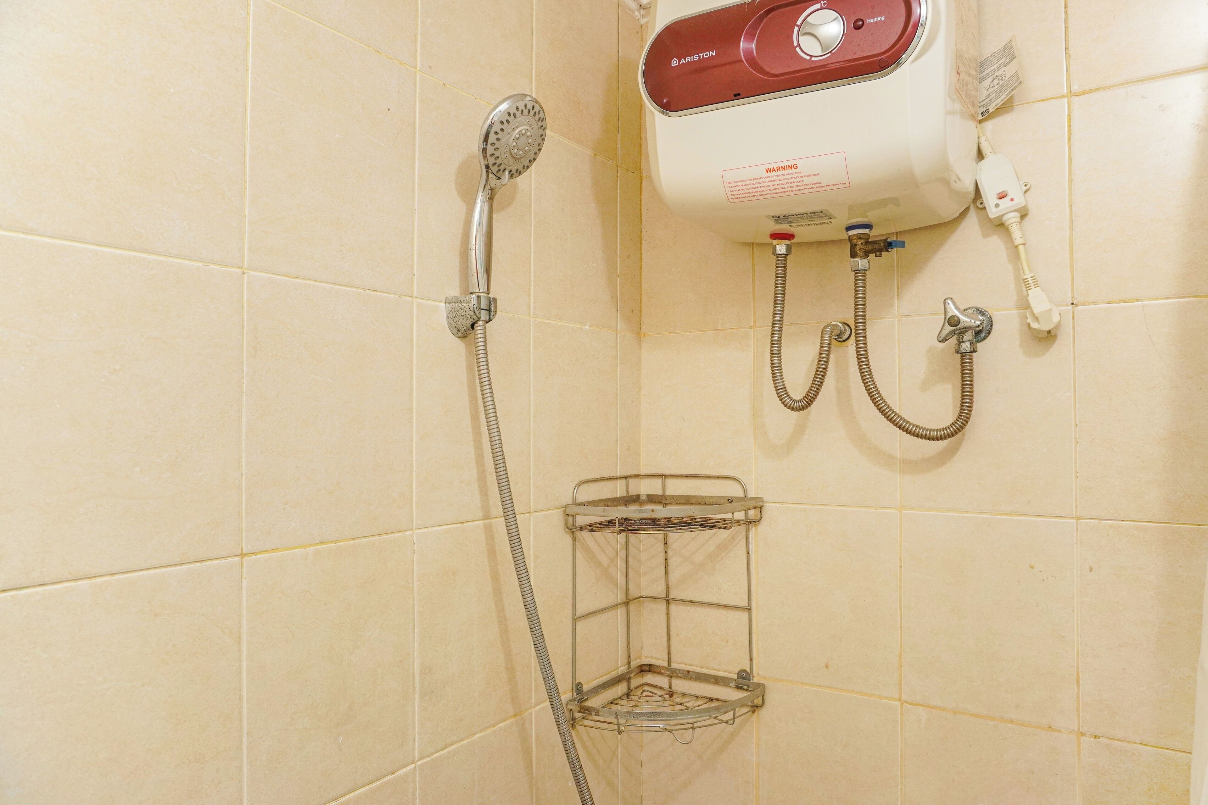 Zona de ducha con ducha de mano, calentador de pared y lavamanos en esquina con estante metálico.