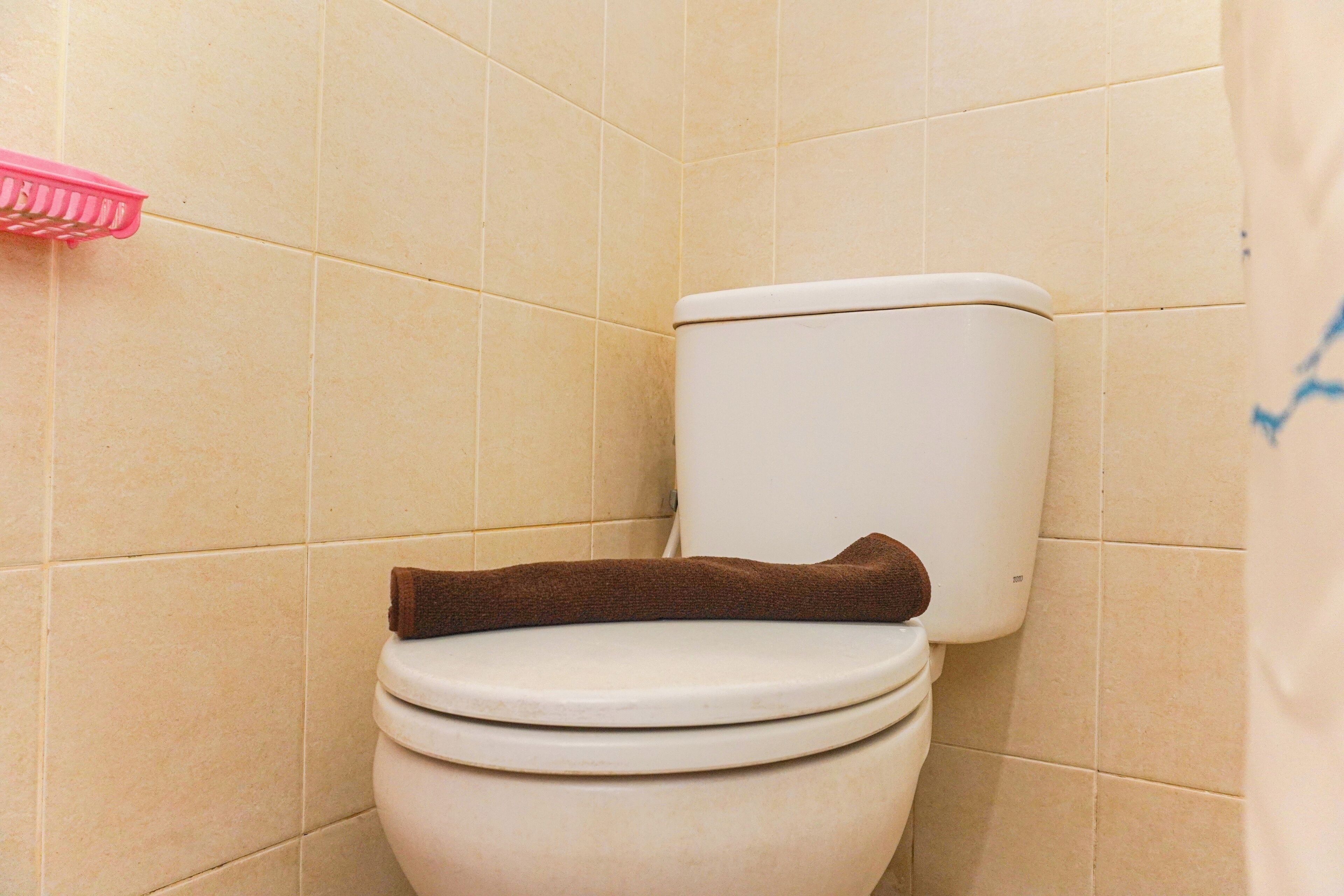 Un inodoro con una toalla marrón sobre el asiento, paredes de azulejos color beige y un cepillo de limpieza rosa a la izquierda.