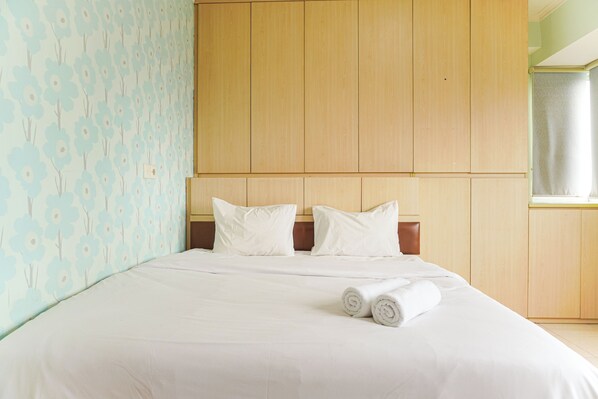 A neatly made bed with white linens and two rolled towels, a wooden headboard, and a floral-patterned wall. - Cozy Living 1Br Maple Park Sunter Apartment (Jakarta)
