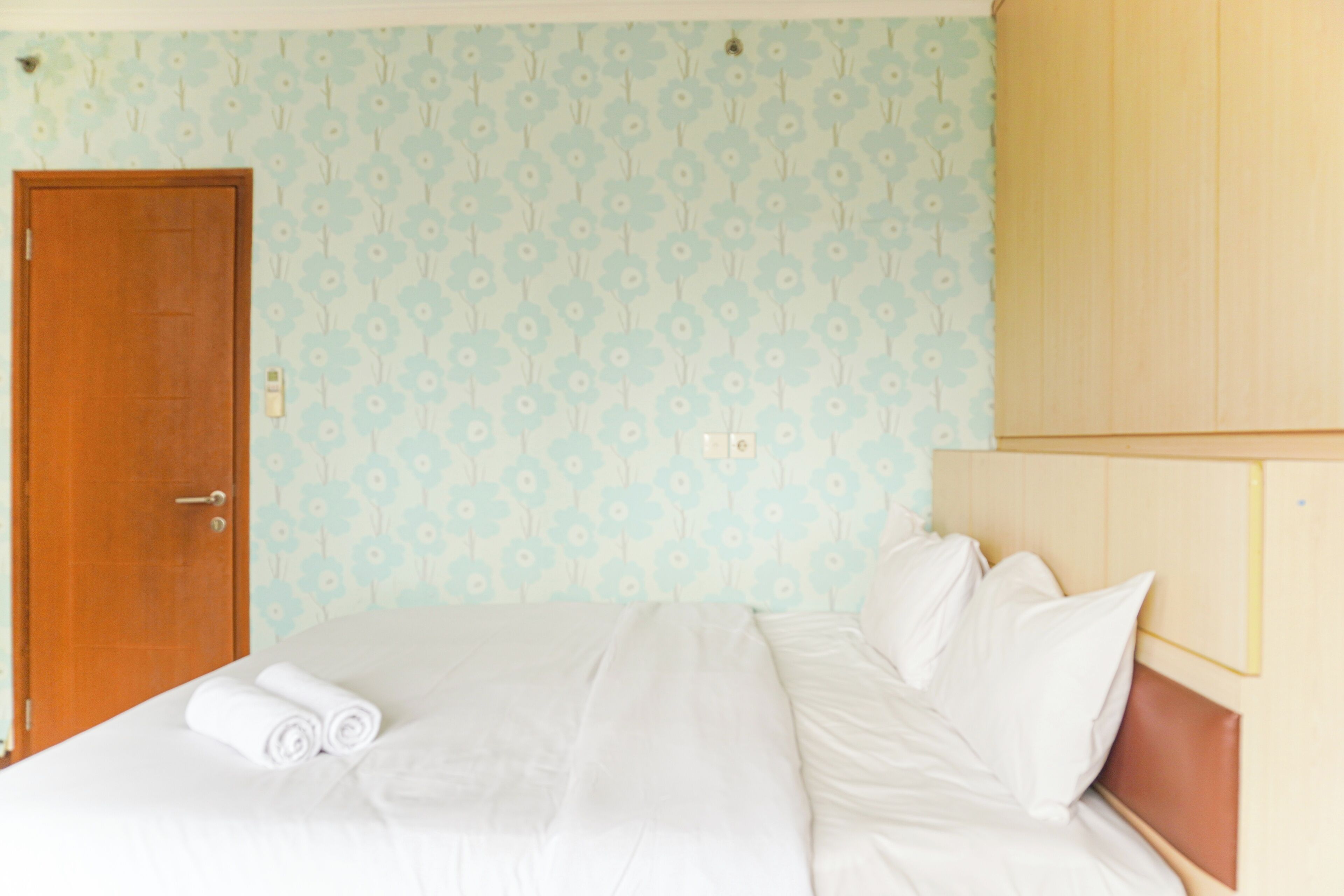 A neatly made bed with white linens and pillows, a wooden door, and a light blue floral wallpaper.