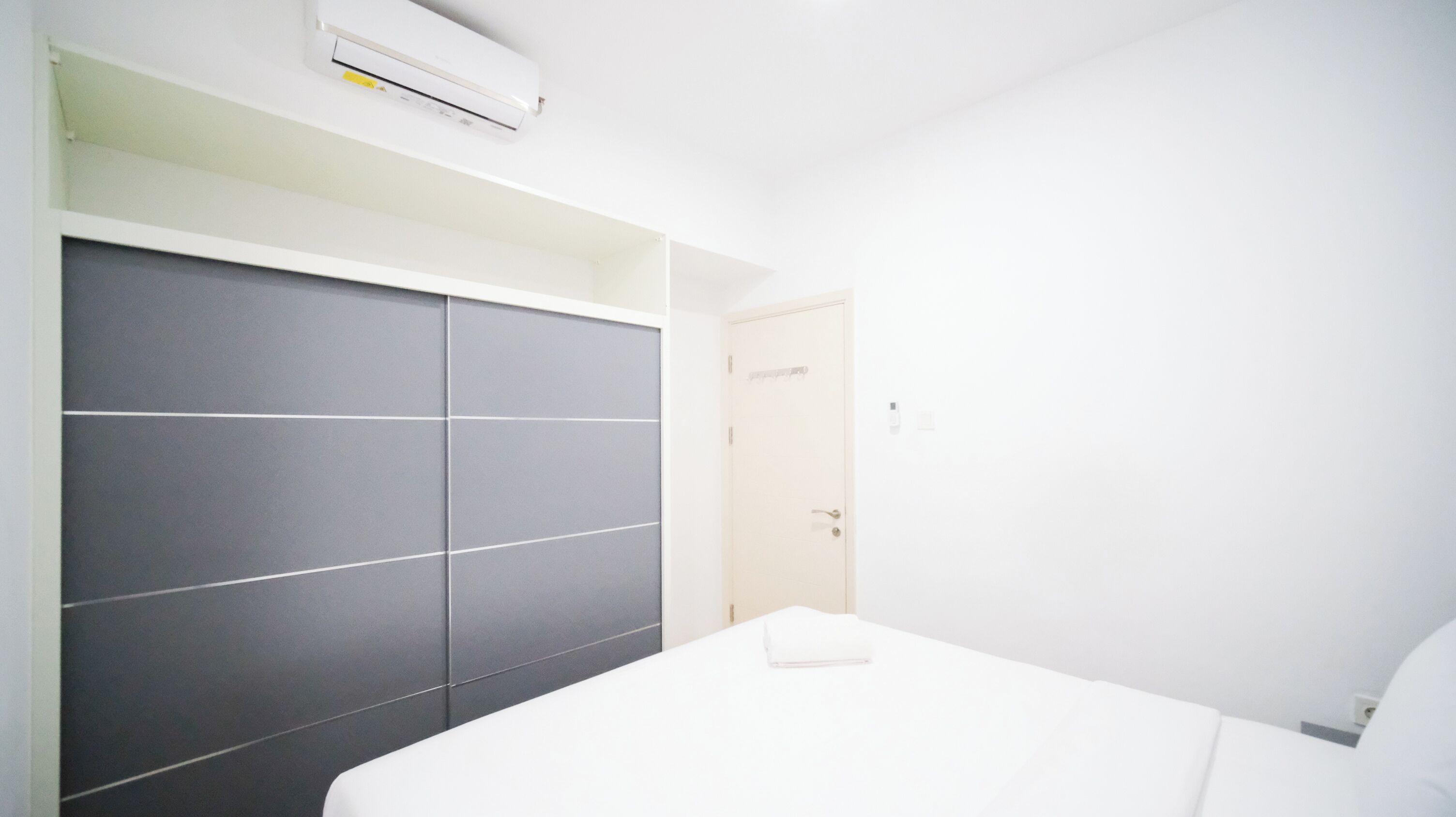 A modern bedroom with a large dark-colored wardrobe, a white bed, and an air conditioning unit mounted on the wall.
