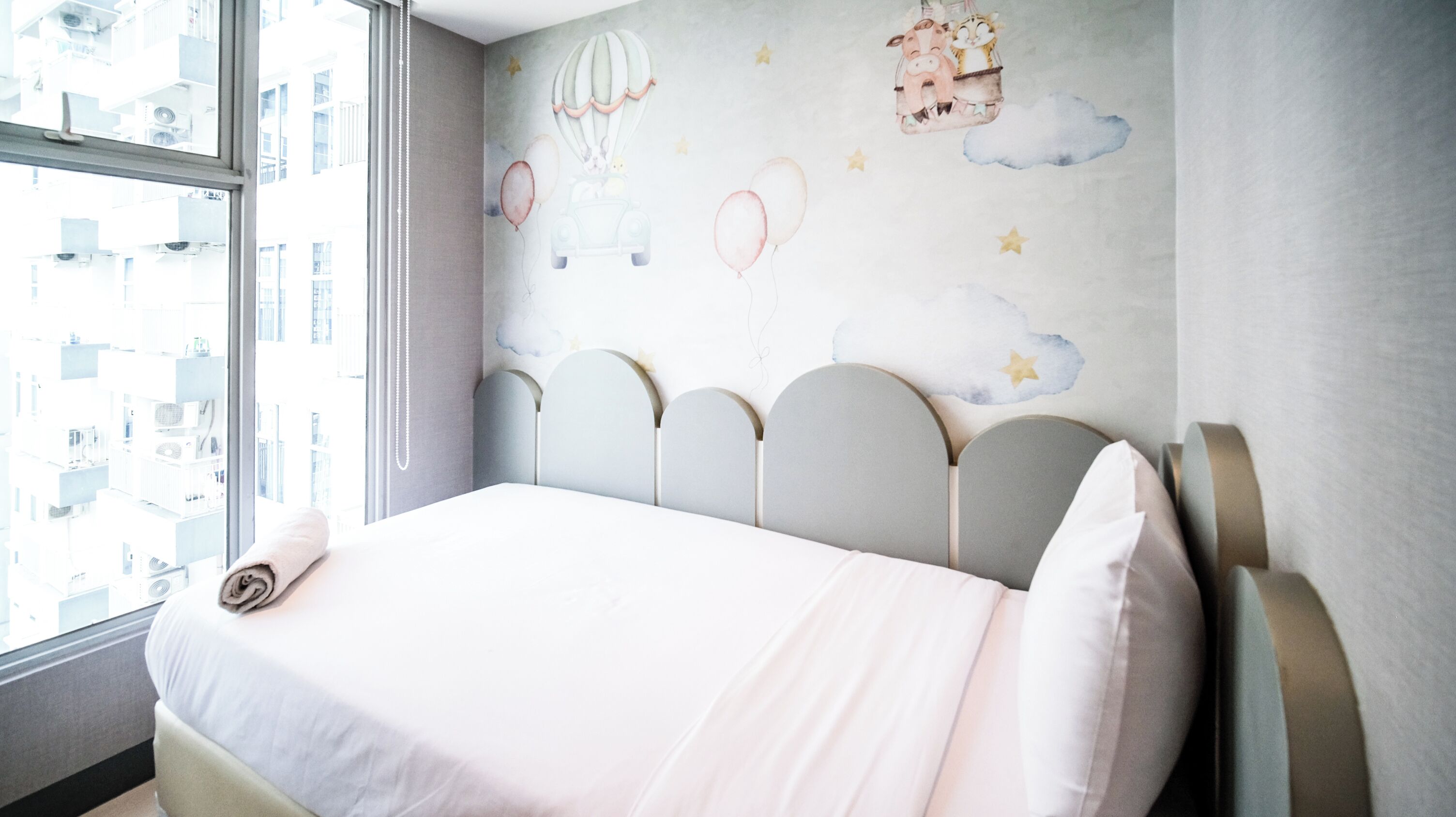 Une chambre d’hôtel avec un lit, une vue sur les immeubles par la fenêtre et un mur décoré d’une fresque pour enfants.