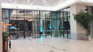 A modern hotel lobby with glass doors, a reception desk, and a large potted plant.