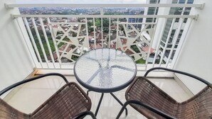 A balcony with a glass table and wicker chairs, overlooking a cityscape with buildings and greenery.