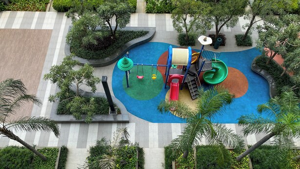 Eine Vogelperspektive zeigt einen Spielplatz mit Rutsche, Schaukeln und einer Kletterkonstruktion, umgeben von viel Grün und einem Pflasterweg.