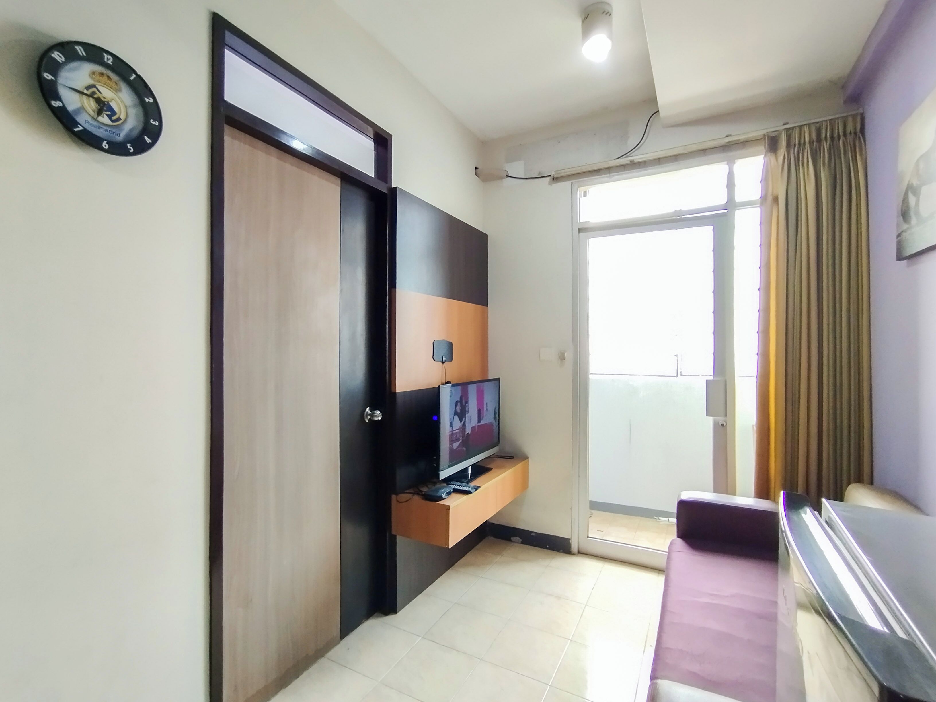 A room with a wooden wardrobe, a television on a stand, a small shelf, and a door leading to a balcony.