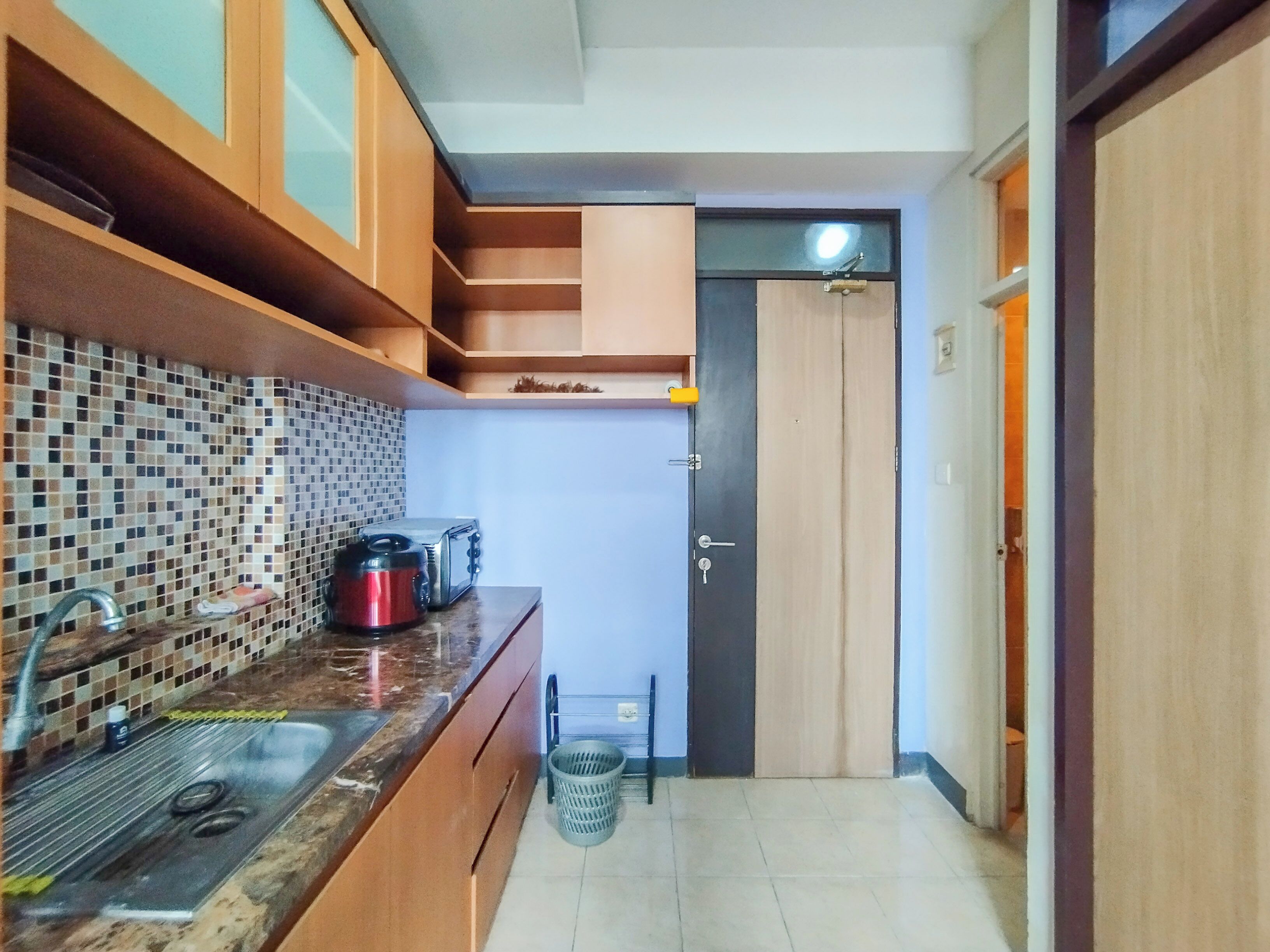 A compact kitchen with a tiled backsplash, a sink, a stove, and a toaster. There is a door leading to another room and a trash bin.