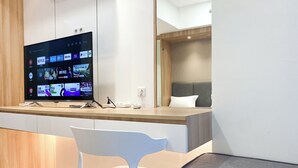 A modern hotel room with a wooden desk, a flat-screen TV displaying various channels, a white chair, and a bed with a grey headboard. - Comfort Living Studio Pollux Chadstone Apartment (Cikarang)