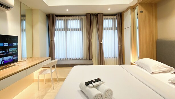A hotel room with a bed, a desk with a computer, a chair, and a TV displaying various channels. - Comfort Living Studio Pollux Chadstone Apartment (Cikarang)