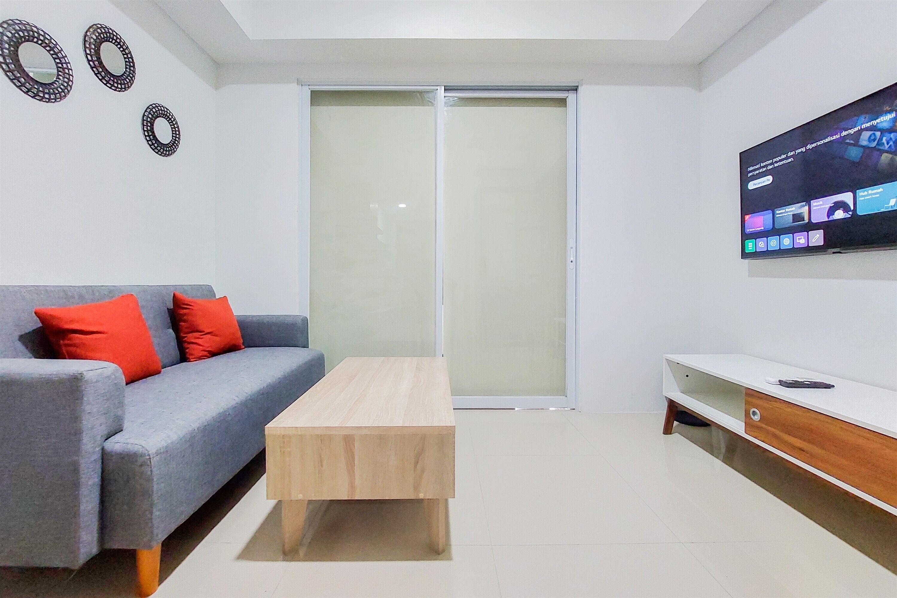 Un salon moderne avec un canapé gris, des coussins rouges, une table basse en bois, un téléviseur fixé au mur et une porte-fenêtre coulissante.