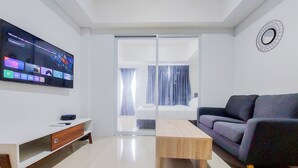 A modern living room with a flat-screen TV, a sofa, a coffee table, and a sliding glass door leading to a balcony. - Homey And Simply Look 1Br Tamansari Bintaro Mansion Apartment (South Tangerang)