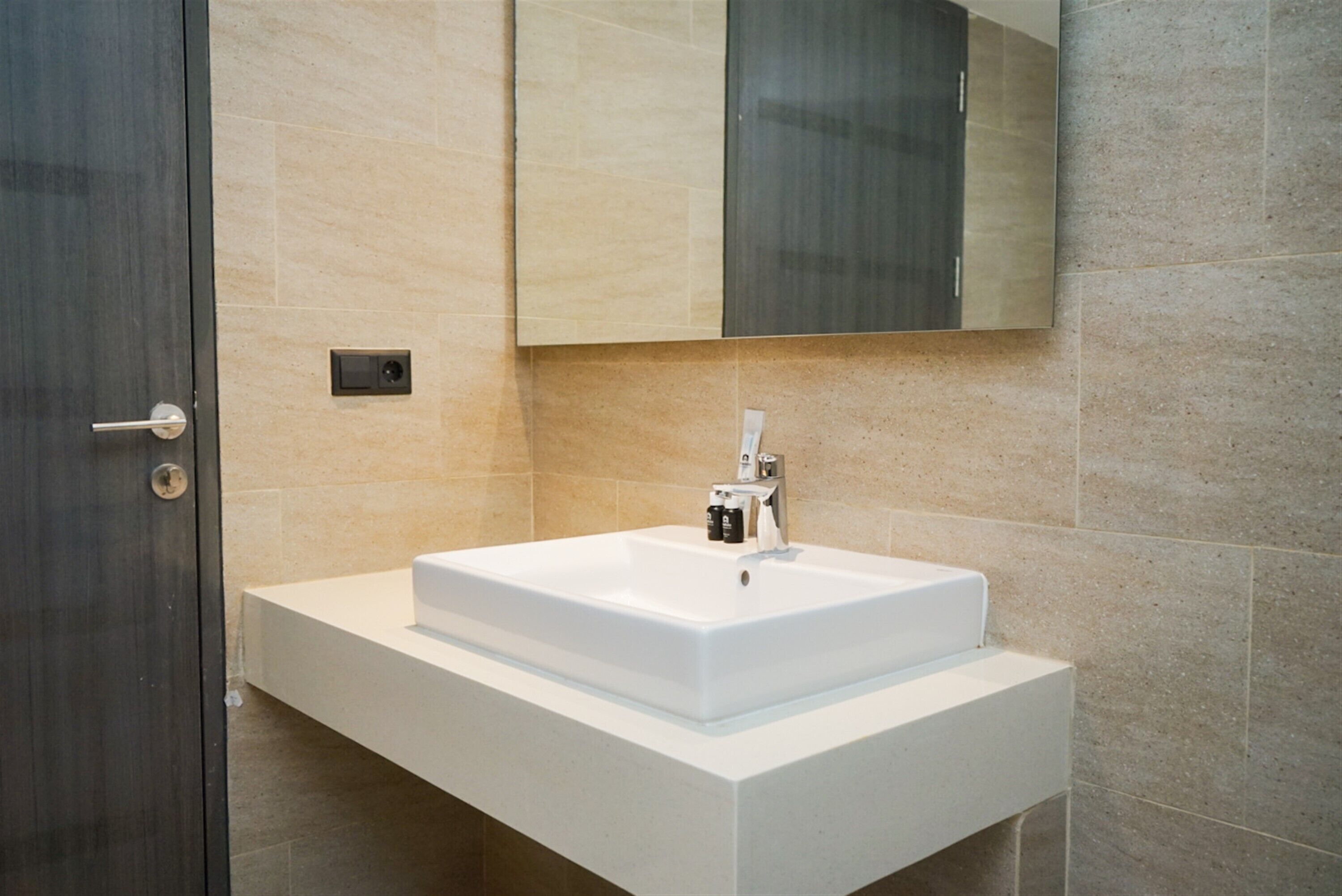 A modern bathroom with a white sink, a dark door, and beige tiled walls.