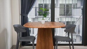 Une table ronde en bois, sur laquelle sont posés un vase blanc et une plante, est flanquée de deux chaises grises. La pièce est dotée de grandes fenêtres et offre une vue sur un immeuble.