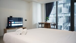 Une chambre d’hôtel avec un lit, un téléviseur affichant l’interface d’un service de diffusion en continu, une petite table avec des chaises et une vue sur un immeuble à travers de grandes fenêtres.