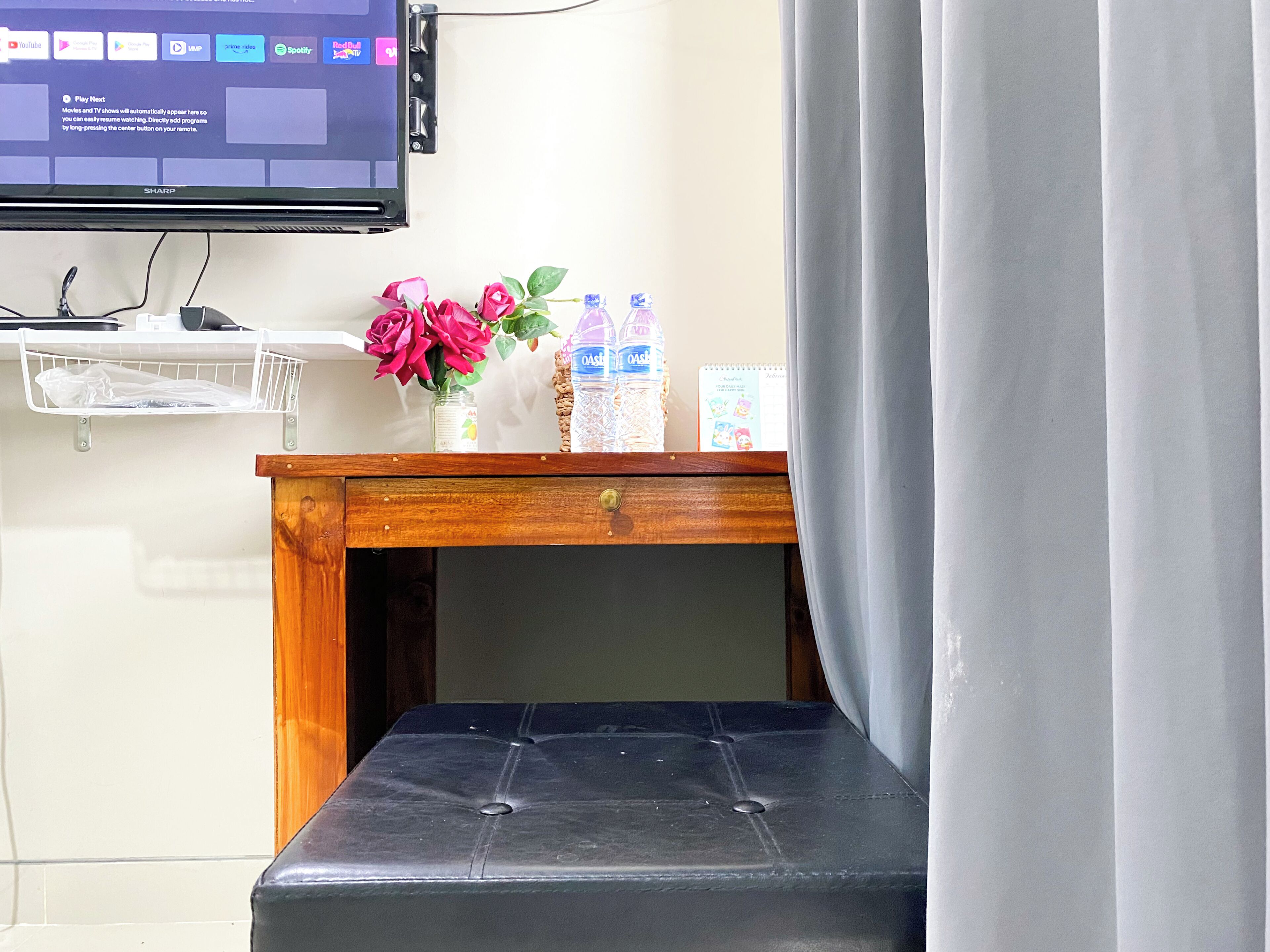 Una taula de fusta petita amb un tamboret negre amb coixí al davant, un televisor de pantalla plana muntat a la paret, i una estant amb un ram de flors i ampolles d'aigua.