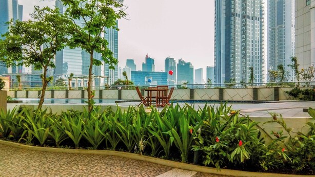 A rooftop garden with a table and chairs, surrounded by lush greenery and tall buildings in the background. - Brand New And Simply 1Br At The Wave Kuningan Apartment (Jakarta)