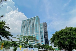 Un edificio moderno de gran altura con fachada de vidrio, rodeado de abundante vegetación y bajo un cielo parcialmente nublado.