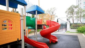 Une aire de jeux avec un toboggan rouge, une structure d’escalade verte et une cabane jaune.