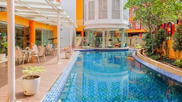 A swimming pool with a tiled edge, surrounded by a covered patio area with chairs and tables, and a building with colorful exterior walls in the background.