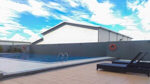 Ein Schwimmbecken mit gefliestem Sonnendeck, Liegestühlen und einem Gebäude im Hintergrund.