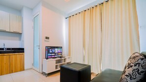 A modern living room with a black leather sofa, a small black ottoman, a flat-screen TV, and a kitchen area with wooden cabinets and a black countertop. - Best Choice And Restful 1Br Citralake Suites Apartment (Jakarta)