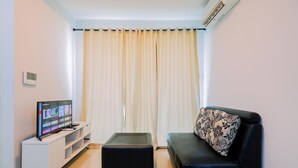 A living room with a flat-screen TV on a white stand, a black ottoman, a black sofa with a floral pillow, and a large window with beige curtains. - Best Choice And Restful 1Br Citralake Suites Apartment (Jakarta)