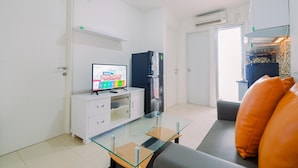 A modern living room with a glass-top coffee table, a white TV stand with a TV displaying a news channel, a black refrigerator, and a grey sofa with orange cushions.