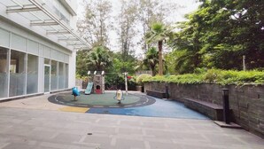 A playground area with a slide and swings, surrounded by a building with large windows and a paved walkway. - Modern And Comfy Studio At Grand Sungkono Lagoon Apartment (Surabaya)