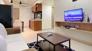 Een moderne woonkamer met een bank, een salontafel en een tv waarop staat 'BEFORE I MET YOU'. Er is een keuken met houten kastjes en een magnetron.