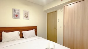 Een net opgemaakt bed met witte lakens en twee opgerolde handdoeken, een houten hoofdbord en twee kunstwerken met bladmotieven aan de muur.