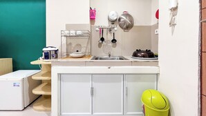 A compact kitchen with a sink, a small refrigerator, and a microwave. There is a wooden shelf with dishes and a yellow bucket.