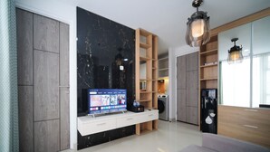 A modern living room with a flat-screen TV on a white console, a black marble accent wall, wooden shelving, and a black refrigerator. - Comfy 2Br At Taman Melati Surabaya Apartment (Surabaya)