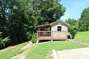 A small cabin with a wooden deck and a covered porch, surrounded by lush greenery and trees.