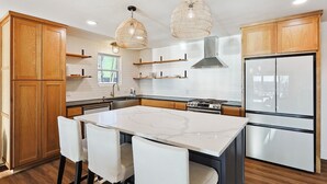 Una cocina moderna con armarios de madera, una isla de mármol, sillas blancas e iluminación colgante.