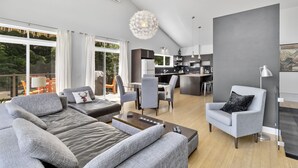 A modern living room with a grey sofa, a coffee table, and a grey armchair. There is a dining area with a table and chairs, and a kitchen in the background.