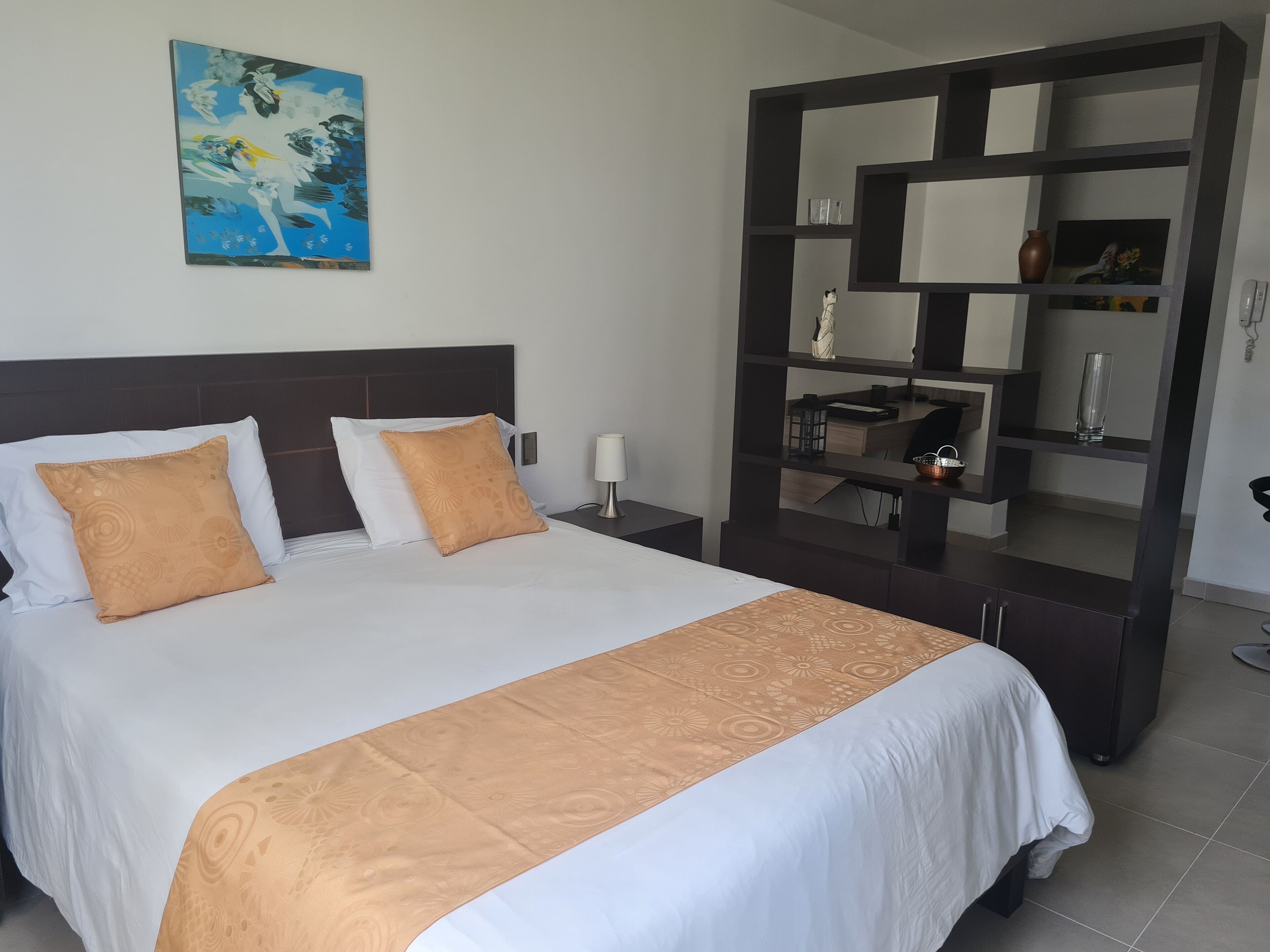 A hotel room with a large bed, two pillows, a nightstand, a lamp, a mirror, and a painting on the wall.
