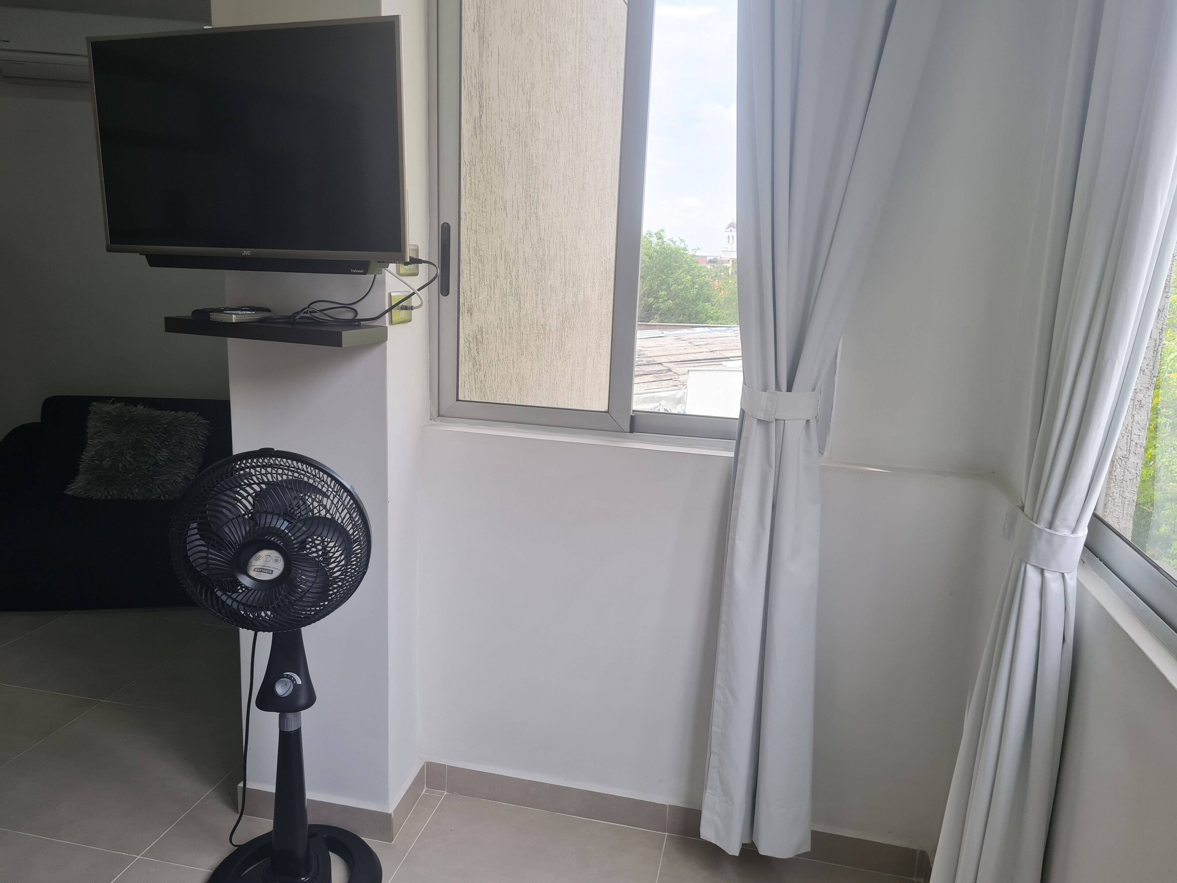 Een kamer met een wandgemonteerde flatscreen-tv, een ventilator, een raam met witte gordijnen en een betegelde vloer.